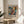 May include: Framed abstract art poster with vibrant swirling colors in blues, oranges, and yellows displayed on a round wooden side table next to a beige upholstered chair. Natural sunlight streams across white walls, with green plant leaves visible in the corner. The poster features bold text reading "POLLOCK" and exhibition details below the colorful abstract artwork.