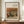 May include: Framed art print featuring Renoir's still life painting of a large striped melon and peaches in warm earth tones, displayed on wooden sideboard next to white ceramic vase and stacked books in elegant room with cream walls and white molding.