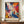 May include: Abstract expressionist poster with dynamic swirling paint splatters in vibrant blues, oranges, reds, and purples against white background, displayed in thin brown frame on wooden sideboard next to white ceramic vase and stacked books in bright room with cream walls and white trim molding.