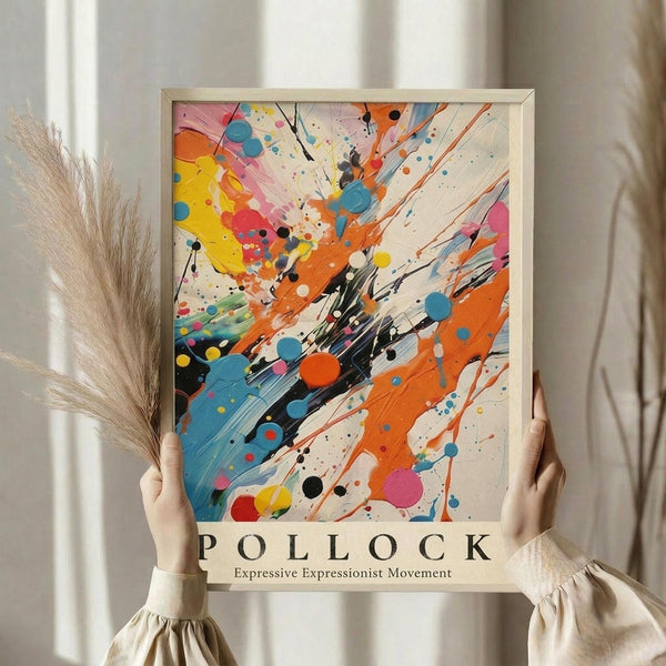 May include: Person holding framed abstract expressionist poster with colorful paint splatters and drips in blue, orange, yellow, pink and black on cream background, standing near dried pampas grass against white curtained window