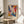 May include: Abstract expressionist art print in thin wooden frame displaying vibrant swirling colors including blue, orange, pink, purple, and white paint strokes. The framed poster sits on a round wooden side table next to a beige upholstered chair, with a plant casting shadows on white walls in natural sunlight.