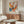 May include: Colorful abstract art poster with vibrant paint splatters in orange, blue, yellow, and pink displayed above a mid-century modern walnut credenza. The framed artwork features dynamic brushstrokes and drips against a cream background. Scene includes a white curved chair, glass oval coffee table, and modern table lamp in a minimalist living room with white walls and natural lighting.