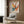 May include: Abstract expressionist art poster with vibrant paint splatters in orange, blue, yellow, pink and black on cream background, displayed in light wood frame on wooden wall panel. Modern beige armchair with geometric patterned pillow sits below artwork, alongside brass floor lamp and dark round side table with open book.