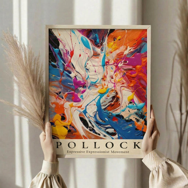 May include: Person in cream sweater holding framed abstract art print with vibrant paint splatters and swirls in blues, oranges, purples, and yellows. Dried pampas grass visible to left, soft beige curtains in background. Poster features bold gestural brushstrokes and drip painting technique with "POLLOCK" text below artwork.