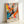 May include: Framed abstract expressionist style poster with vibrant paint splatters and drips in orange, blue, yellow, pink, and black against white background. Features dynamic brushstrokes and color bursts radiating from center. Text reads "POLLOCK Expressive Expressionist Movement" at bottom. Light wooden frame leans against white wall, showing natural lighting and shadows.