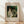 May include: Framed vintage exhibition poster featuring Pierre-Auguste Renoir's "La Promenade" from 1870, showing a woman in white dress and man in dark clothing walking through dappled forest sunlight. The cream-colored poster with elegant typography sits against white fabric backdrop on light wood flooring, with soft natural lighting creating diagonal shadows.