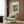 May include: Vintage Renoir art print in cream matted frame hanging on wood-paneled wall above beige upholstered armchair with geometric patterned pillow. The impressionist painting depicts figures near water with lush green foliage. Modern reading corner setup includes brass floor lamp and dark round side table with open book. Neutral tones throughout with natural lighting from nearby window.