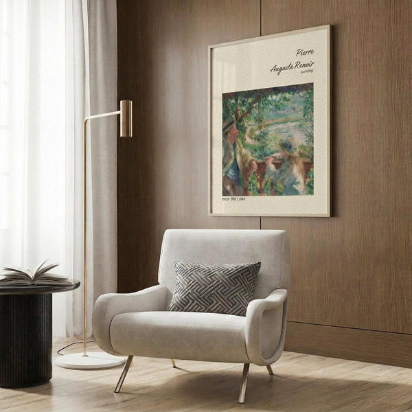 May include: Vintage Renoir art print in cream matted frame hanging on wood-paneled wall above beige upholstered armchair with geometric patterned pillow. The impressionist painting depicts figures near water with lush green foliage. Modern reading corner setup includes brass floor lamp and dark round side table with open book. Neutral tones throughout with natural lighting from nearby window.