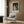 May include: A framed poster featuring an impressionist painting of a man and woman in 19th-century dress walking through a lush green forest. The woman wears a flowing white dress and the scene has soft, dappled lighting. The poster displays "PIERRE AUGUSTE" and "exhibition" text below the artwork. Shown in a modern living room setting with a beige upholstered armchair, geometric patterned pillow, brass floor lamp, and wood-paneled wall.