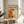 May include: Framed art poster featuring Jackson Pollock's abstract expressionist painting in vibrant orange, yellow, black and white paint splatters and drips, displayed on a light wood round side table next to a beige upholstered chair with green plant leaves visible, natural sunlight casting shadows across white walls in a minimalist modern living room setting.