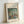 May include: Framed art print in light wooden frame leaning against white wall. Features Pierre-Auguste Renoir impressionist painting showing figures by lakeside with lush green foliage and soft brushstrokes in blues, greens, and earth tones. Print has cream mat border with artist name and title text. Natural lighting creates gentle shadows on wall.