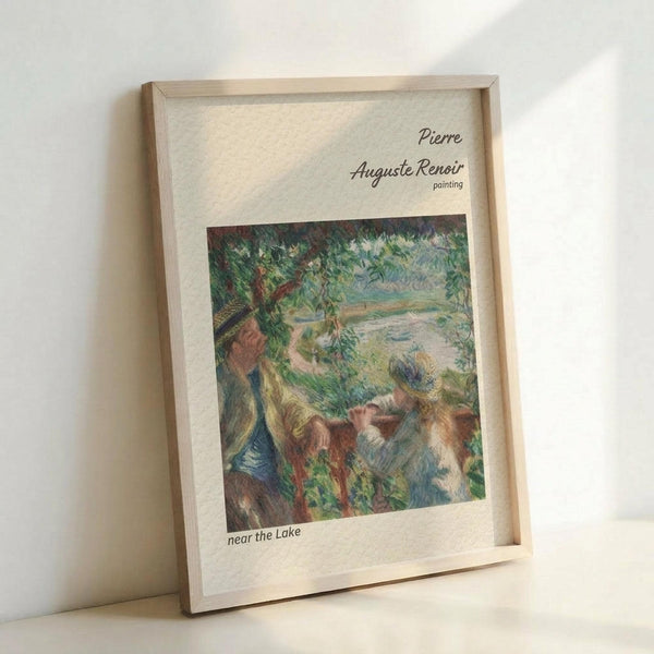 May include: Framed art print in light wooden frame leaning against white wall. Features Pierre-Auguste Renoir impressionist painting showing figures by lakeside with lush green foliage and soft brushstrokes in blues, greens, and earth tones. Print has cream mat border with artist name and title text. Natural lighting creates gentle shadows on wall.