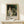 May include: Framed exhibition poster featuring Renoir's impressionist painting of a couple in a forest setting - woman in flowing white dress with blue sash, bearded man in dark clothing and cap. Poster displays "PIERRE AUGUSTE exhibition" text with "1870" and "La Promenade" details. Natural wood frame sits on light wooden surface next to small white wrapped gift with twine and greenery against cream wall.