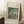 May include: Framed art print of an impressionist landscape painting leaning against white wall corner. The artwork shows figures near water with lush green foliage and soft brushstrokes in blues, greens, and earth tones. Cream matting surrounds the image with artist attribution text. A coffee cup sits on wooden block nearby, with textured cream rug and beige throw blanket visible.