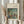 May include: Person in beige clothing holding framed reproduction of Impressionist painting showing figures near water surrounded by lush greenery in soft blues, greens, and earth tones. Light wood frame with cream matting against neutral curtained background. Dried wheat stems visible on left side.