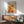 May include: Abstract expressionist poster featuring vibrant orange, white and black paint splatters and gestural brushstrokes, displayed in thin wooden frame above mid-century modern credenza with white ceramic table lamp. Sage green upholstered armchair with wooden frame sits nearby on hardwood floor. Dramatic sunlight creates angular shadows across white wall, highlighting the artwork's dynamic composition and energetic paint application.