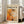 May include: Framed abstract art poster with orange, white, and black paint splatters in Jackson Pollock's drip painting style, displayed on wooden floor against white walls next to beige upholstered chair. Sunlight streams through tall windows creating soft shadows. The artwork features bold text reading "POLLOCK" and "CHAOS AND CREATIVITY 1912-1956" above dynamic brushstrokes and paint drips in warm autumn tones.
