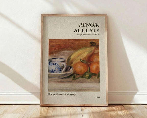 May include: Framed art print leaning against white wall on light wooden floor. The print features a Renoir still life painting showing oranges, bananas, and a blue and white porcelain teacup on a wooden surface. Text reads "RENOIR AUGUSTE" at top with "Oranges, bananas and teacup" and "1908" below the artwork. Natural wood frame with beige matting, soft window light casting shadows across white fabric backdrop.