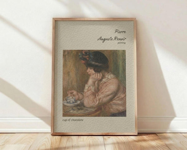 May include: Framed art print leaning against white wall on light wood floor. Wooden frame contains reproduction of Impressionist painting showing woman in dark feathered hat and pink dress drinking from small cup, rendered in soft earth tones. Text above reads "Pierre Auguste Renoir painting" and below "cup of chocolate" on cream textured background.