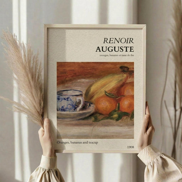 May include: Person in cream ruffled sleeves holding framed art print featuring Renoir's still life painting of oranges, bananas, and blue patterned teacup on wooden surface. Light wood frame against beige textured background. Dried wheat stalks visible on left side. Soft natural lighting creates warm, minimalist aesthetic.
