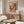 May include: Abstract art exhibition poster with warm orange, red and yellow paint splashes displayed above wooden sideboard in modern living room. White curved chair and glass coffee table visible. Poster features bold "POLLOCK" text and exhibition dates, shown in thin frame against neutral wall.