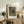 May include: Framed reproduction of an impressionist oil painting showing colorful wildflowers in a brown ceramic vase, displayed on a rustic wooden sideboard beside books, a potted fern, ceramic vessels, and a table lamp in a cozy living room with neutral walls and upholstered furniture.