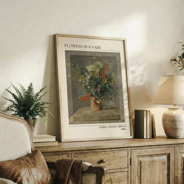 May include: Framed reproduction of an impressionist oil painting showing colorful wildflowers in a brown ceramic vase, displayed on a rustic wooden sideboard beside books, a potted fern, ceramic vessels, and a table lamp in a cozy living room with neutral walls and upholstered furniture.