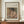 May include: Framed vintage art print showing woman in dark hat and pink dress drinking from teacup, displayed on wooden sideboard next to white ceramic vase and stacked books against cream walls with white trim molding