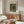 May include: Interior scene showing a framed vintage art poster of wildflowers in a terracotta vase mounted on white wall above mid-century walnut credenza. Modern white sculptural chair, glass coffee table, and minimalist table lamps complete the neutral-toned living space with natural lighting from window.