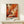 May include: Framed abstract expressionist artwork in warm orange, red, and black paint splatters and brushstrokes, displayed on white wooden console table against cream wall. Small white gift box with evergreen sprig sits beside the light wood frame.