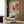 May include: Abstract expressionist exhibition poster featuring vibrant orange, red, and yellow paint splatters with "POLLOCK" text and exhibition dates, displayed in cream frame on wood-paneled wall above beige upholstered chair with geometric patterned pillow, brass floor lamp, and dark round side table with open book nearby.