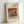 May include: Framed art print leaning against white wall showing Renoir still life painting of oranges, bananas, and blue and white patterned teacup on wooden surface. Print has cream colored mat with French and English text, housed in light wood frame. Warm lighting creates soft shadows on wall.
