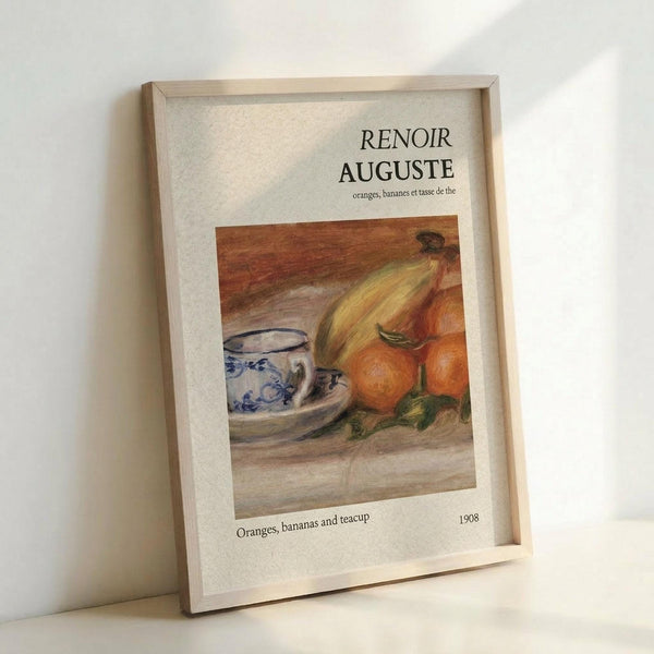 May include: Framed art print leaning against white wall showing Renoir still life painting of oranges, bananas, and blue and white patterned teacup on wooden surface. Print has cream colored mat with French and English text, housed in light wood frame. Warm lighting creates soft shadows on wall.
