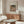 May include: Framed Renoir reproduction print showing woman in pink dress and dark hat holding teacup, displayed above wooden sideboard in modern living room with cream walls, white curved chair, glass coffee table, and warm natural lighting creating soft shadows.