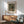 May include: Framed art print of Renoir's impressionist flower painting hanging on white wall above wooden credenza. Print shows colorful wildflowers in earthenware vase against muted background. Modern room setting with mint green upholstered armchair, white ceramic table lamp, and natural sunlight casting shadows across the scene.