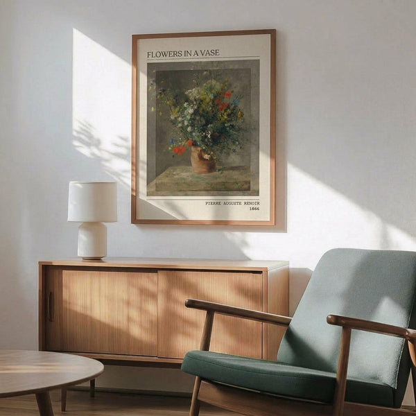 May include: Framed art print of Renoir's impressionist flower painting hanging on white wall above wooden credenza. Print shows colorful wildflowers in earthenware vase against muted background. Modern room setting with mint green upholstered armchair, white ceramic table lamp, and natural sunlight casting shadows across the scene.