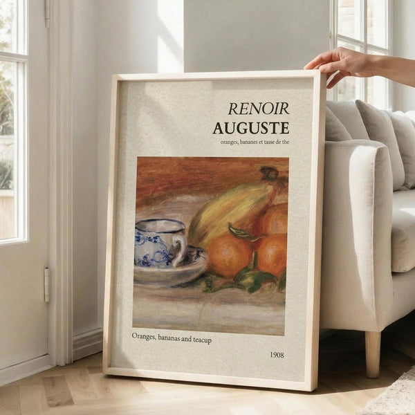 May include: A framed art print featuring a Renoir still life painting of oranges, bananas, and a blue and white patterned teacup, displayed in a bright living room with beige furniture, white walls, and large windows. The poster has cream matting with text above and below the artwork, held by a person's hand against a neutral sofa.