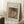 May include: Framed Renoir reproduction showing woman in pink dress and dark feathered hat holding teacup, leaning on hand in contemplative pose. Warm earth tones with impressionist brushwork. Light wooden frame with cream matting displays "Pierre Auguste Renoir painting" and "cup of chocolate" text. Propped against white wall on textured cream rug beside wooden stool with coffee cup, creating cozy room setting with natural window light.