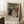 May include: A framed exhibition poster featuring Renoir's impressionist painting of a family group with adults and children in period dress, displayed in a bright room setting. The beige-framed print shows soft, warm tones and classic composition. Scene includes a white coffee cup on wooden surface and cream textured rug, with natural window light creating gentle shadows on white walls.