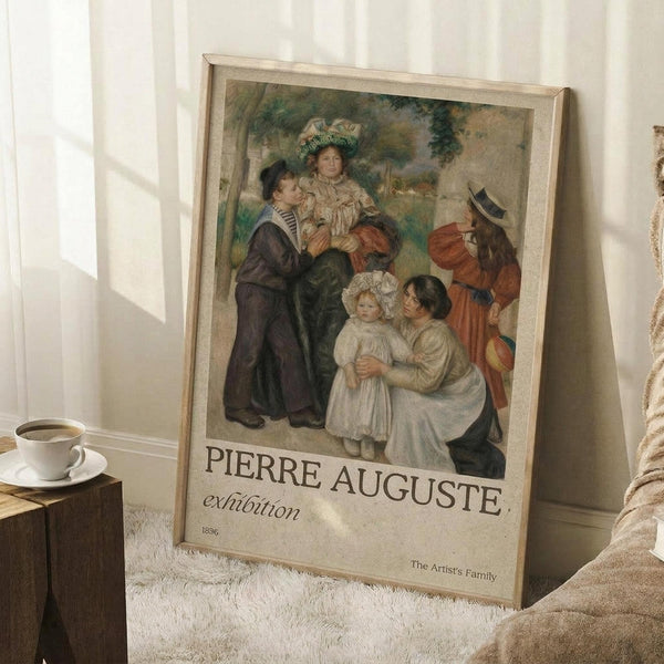 May include: A framed exhibition poster featuring Renoir's impressionist painting of a family group with adults and children in period dress, displayed in a bright room setting. The beige-framed print shows soft, warm tones and classic composition. Scene includes a white coffee cup on wooden surface and cream textured rug, with natural window light creating gentle shadows on white walls.