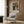 May include: Beige armchair with geometric patterned pillow positioned next to wood paneled wall displaying framed exhibition poster featuring Renoir's colorful family portrait painting. Modern floor lamp with brass finish and circular white base stands nearby. Natural lighting from window with curtains creates warm, contemporary living room setting.