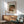 May include: Framed Renoir reproduction poster showing a woman in orange dress with blonde child playing with toys, displayed on white wall above wooden credenza with table lamp, next to mint green mid-century modern armchair with wooden arms in sunlit room with hardwood floors.
