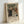 May include: Framed exhibition poster featuring Renoir's 1896 painting of a family group in period clothing - adults and children in white bonnets and dark formal wear against a soft green background. Light wooden frame with cream matting, leaning against white wall. Poster displays "PIERRE AUGUSTE exhibition" text below the artwork with "The Artist's Family" and "1896" details.