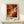 May include: Abstract expressionist artwork in warm orange, red, and black swirling paint strokes displayed in a light wood frame on a white wall. The framed print sits on a pale wooden console table next to a small white gift box with green botanical decoration. The painting features dynamic fluid movements and bold contrasting colors in an energetic composition.