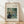 May include: Framed art print of Renoir's impressionist painting showing two figures dining at a restaurant terrace with green lattice work and foliage. The poster features soft brushstrokes in blues, greens, and warm tones, displayed in a light wood frame with cream matting, leaning against white wall on blonde hardwood floor with natural window light creating shadows.