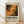 May include: Framed exhibition poster featuring abstract paint splatters in orange, black, and cream colors with dripping and splashing effects. Poster displays "POLLOCK" in large black letters with exhibition dates "MAY 18, 2030 - AUGUST 14, 2030" below. Natural wood frame leans against white wall on light wooden floor with soft shadows cast across the scene.