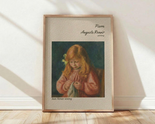 May include: Framed vintage art print featuring a young girl with long reddish-blonde hair in a pink dress, focused on sewing. The reproduction shows Renoir's soft impressionist style with warm tones against a dark green background. Displayed in a natural wood frame with cream matting, leaning against a white wall on light wooden flooring with natural window light creating gentle shadows.