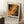 May include: Framed abstract art poster with Jackson Pollock-style paint splatters in autumn colors of orange, yellow, black and cream, leaning against white wall next to wooden side table with coffee cup, positioned on light textured rug with beige throw blanket visible at edge.