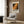 May include: Framed abstract art poster with vibrant orange, black and cream paint splatters hanging on wood-paneled wall above cream upholstered armchair with geometric throw pillow. Modern brass floor lamp and round black side table with open book visible. Warm neutral interior with natural lighting from window.