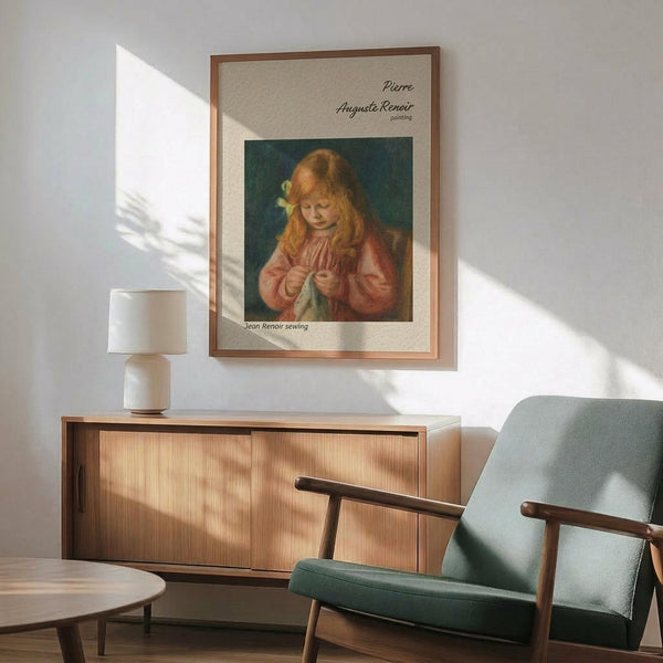 May include: Framed vintage Renoir poster print showing young girl with reddish hair and yellow flower sewing in pink dress against dark green background, displayed on white wall above wooden sideboard with ceramic lamp, next to sage green mid-century modern armchair with wooden frame in sunlit room